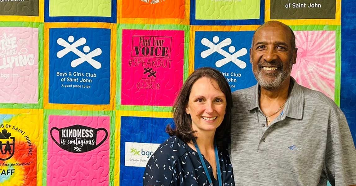 A smiling woman and man standing in front of a colorful quilt featuring "Boys & Girls Club of Saint John" logos.