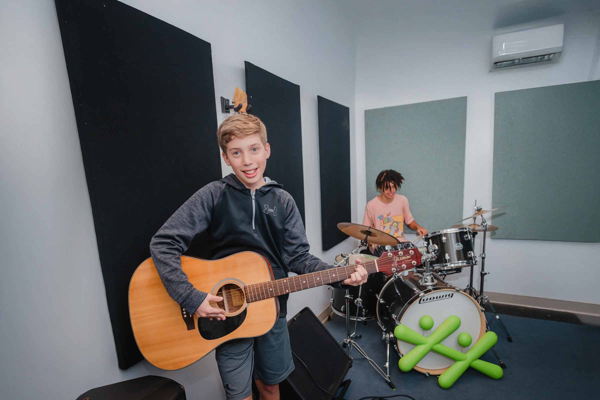 A young boy plays an acoustic guitar while another youth plays the drums in a music room featuring a green BGC logo on the bass drum.