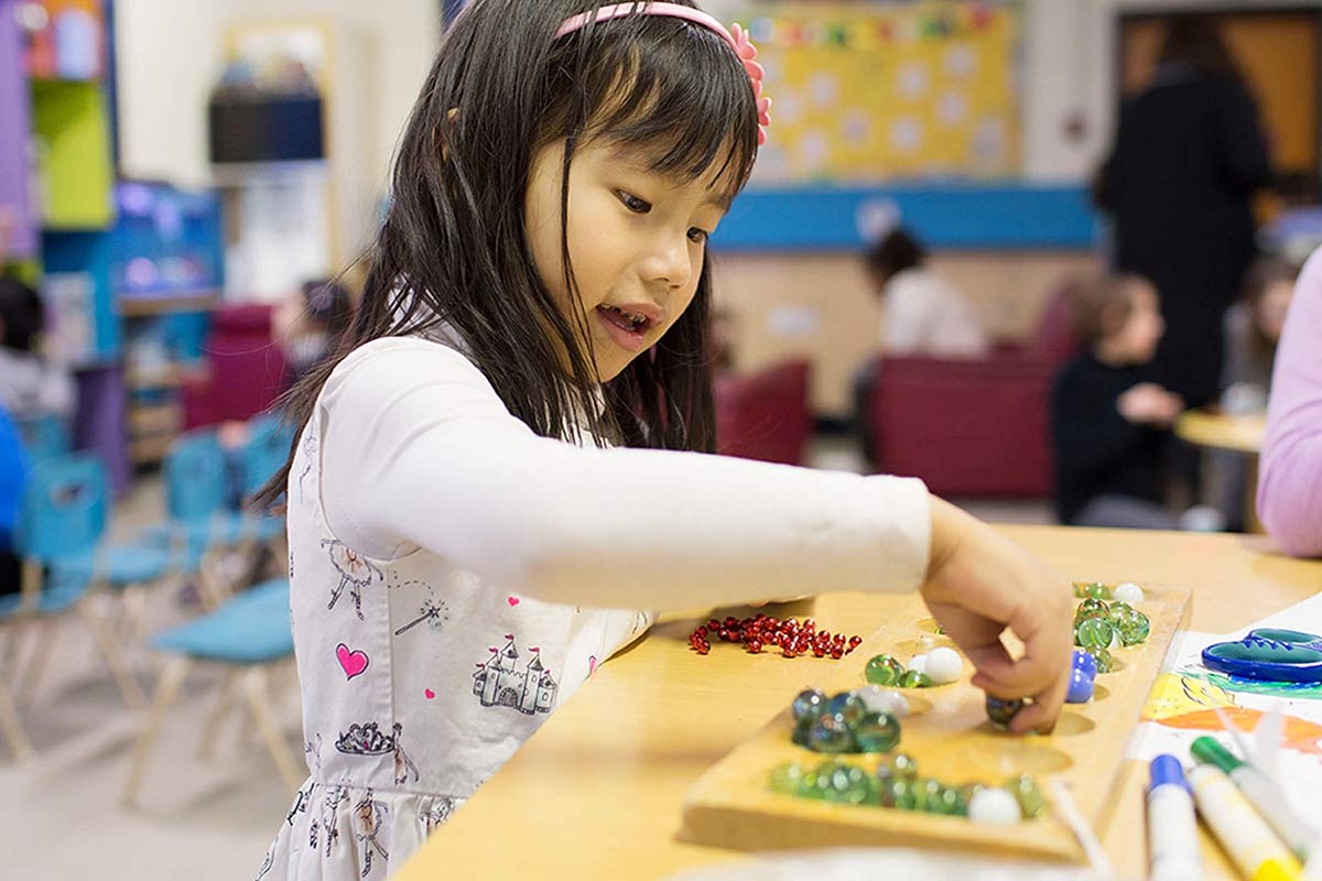 A young girl wearing a patterned white dress plays with glass marbles on a wooden board in a colorful classroom setting.