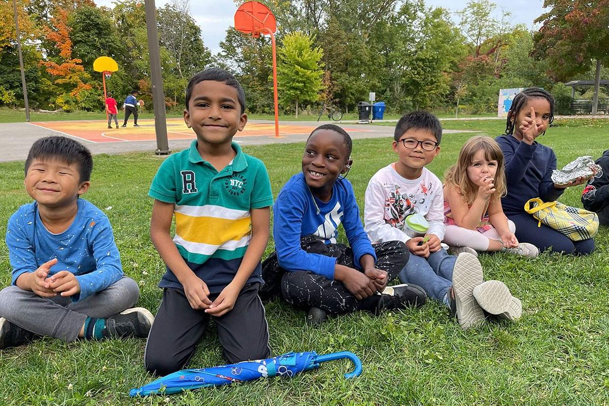 A group of young children sitting on the grass at a park, smiling and enjoying time together near a playground