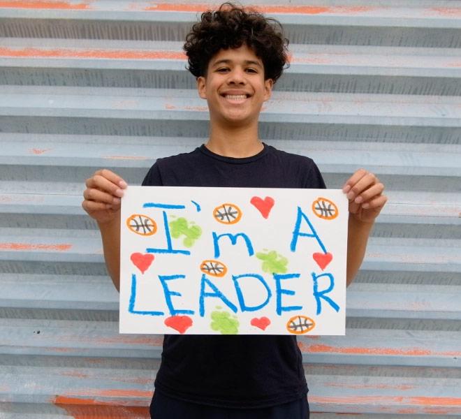 A youth smiling and holding a handmade sign that reads ‘I’m a leader,’ decorated with colorful drawings.