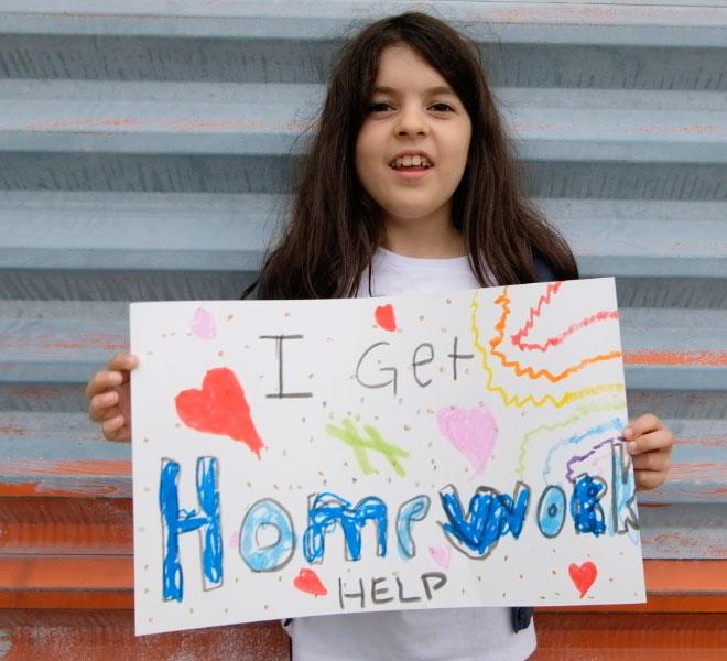 A young girl holds up a colorful, hand-drawn sign that reads "I Get Homework Help" while standing in front of blue steps.