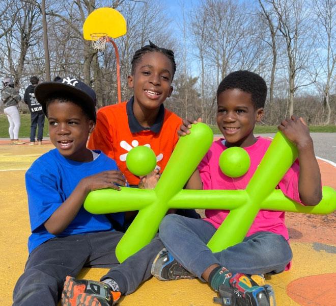 Three children sitting together outdoors at a park, smiling and holding green inflatable shapes during a group activity.
