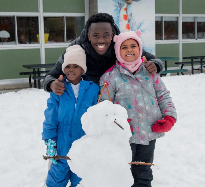 Three children standing together outdoors in winter, smiling beside a snowman.