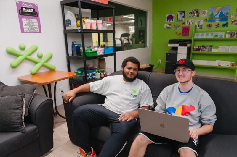Two young men sitting on a couch, one using a laptop in a community center room.