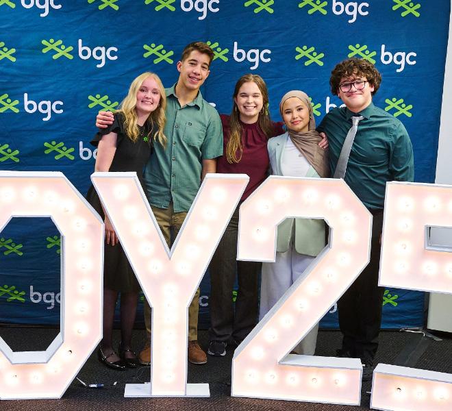 Five smiling young people pose for a group photo behind large illuminated letters in front of a blue backdrop patterned with the BGC logo.