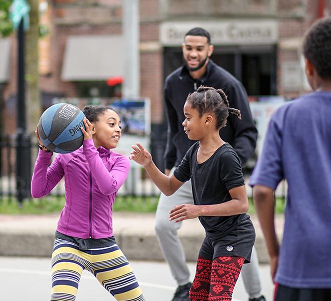 A young girl in a purple jacket holds a basketball while playing outdoors with other children and an adult.