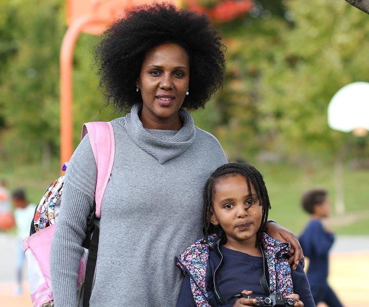A woman with an afro hairstyle stands outdoors with her arm around a young girl who is holding a camera.