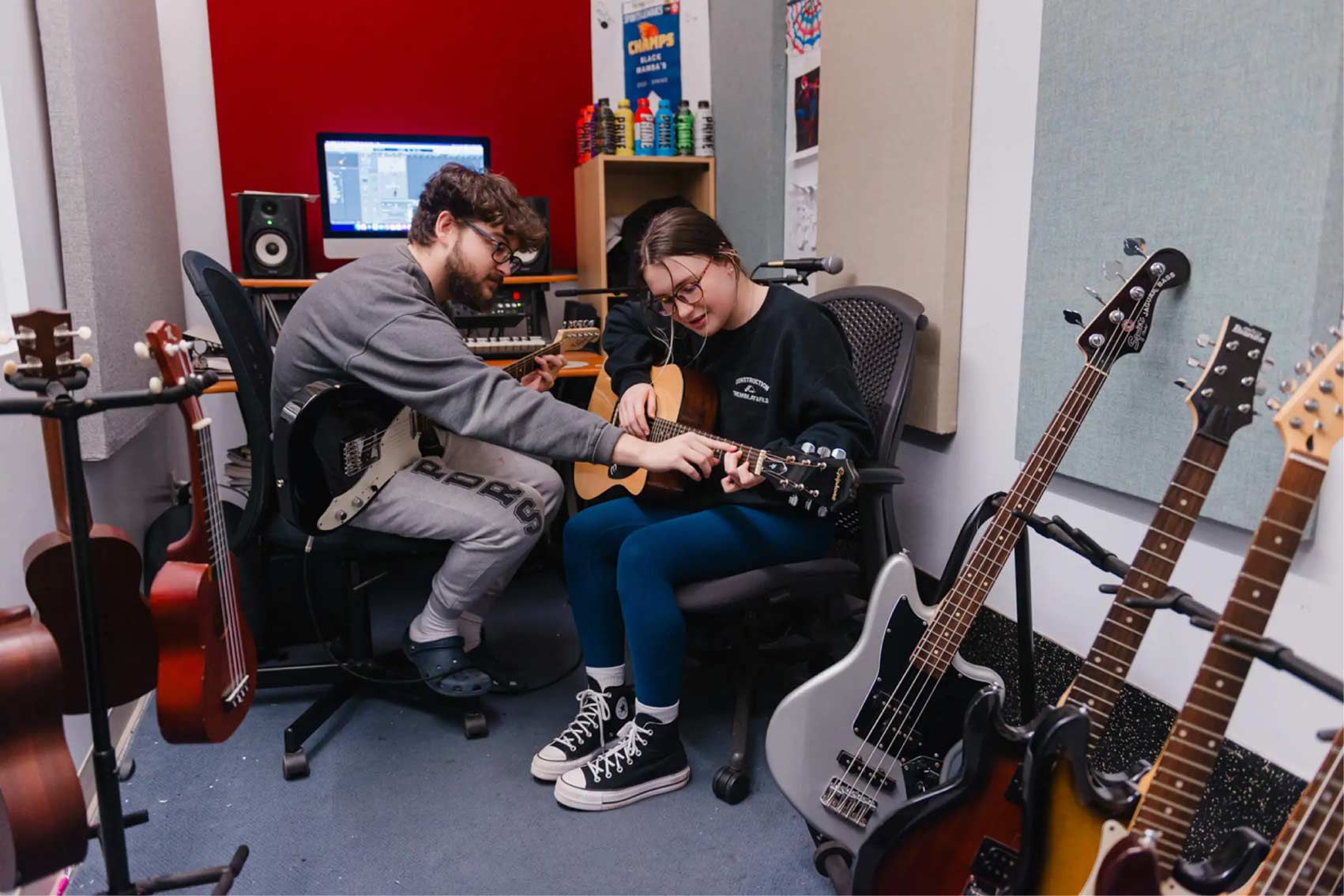 A male youth mentor teaches a young girl how to play the guitar in a recording studio surrounded by musical instruments.