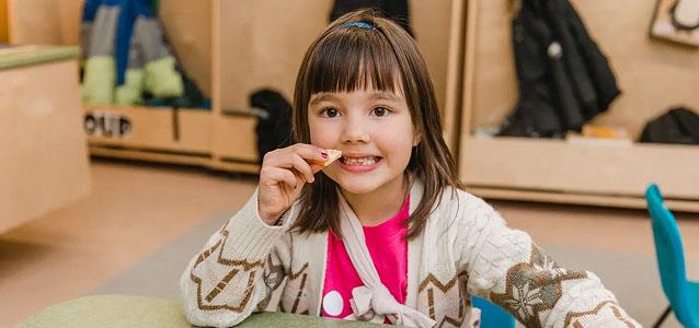 A young girl with brown hair and a pink shirt smiles while eating an orange slice at a table.