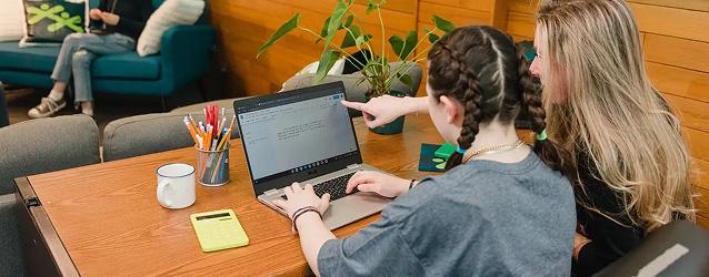 Two young women collaborate on a laptop at a wooden table with a calculator and pencils.