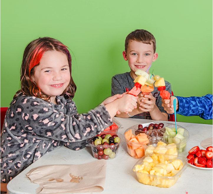 Three smiling children toasting with fruit skewers at a table filled with containers of fresh fruit.