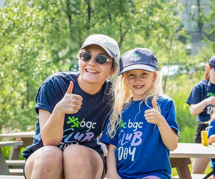 Alternative text: A woman and a young girl both wear blue "BGC Club Day" t-shirts and smile while giving a thumbs up at an outdoor picnic table.