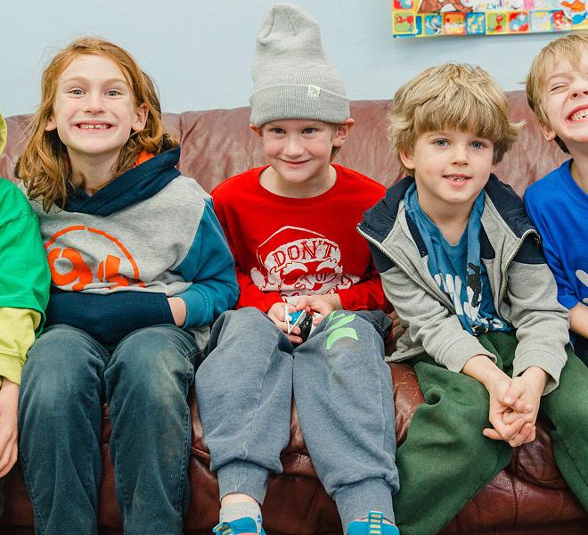 Four young children sit together on a brown leather couch, smiling and making playful faces.