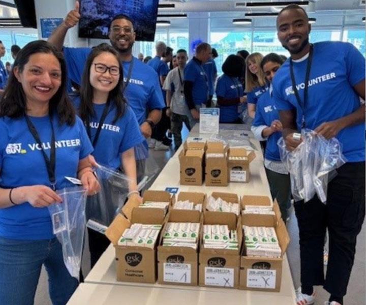 Four volunteers wearing blue "VOLUNTEER" t-shirts smile while packing boxes of consumer healthcare products at a long table.