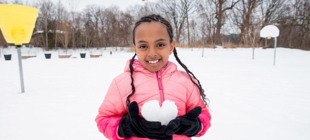 A young girl with braided hair and a pink winter jacket smiles while holding a heart-shaped snowball in her gloved hands. She is standing in a snowy park with a yellow ball toss game and a basketball hoop visible in the background.