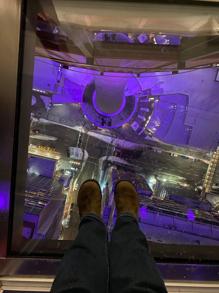 A first-person perspective looking down through a glass floor pane, showing a person's feet in brown boots and jeans dangling over a dramatic drop. Below, the base of a towering structure is lit up in vibrant purple, looking down onto city streets with small cars and buildings far below at night.