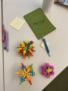 A top-down shot of a white desk with three colorful, interlocking ball-and-joint toy sculptures in the foreground. In the background sits a green notebook branded with the 'bgc Canada' logo, accompanied by a blue pen, sticky notes, and three colored markers on the left side.