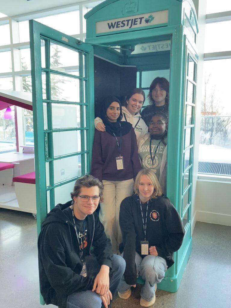 A group of six diverse teenagers smiling for a photo. In the foreground, two are crouching. Behind them, four stand closely inside and in front of an open, teal-colored retro phone booth labeled 'WESTJET'. The background shows a modern, sunlit interior with large windows and other seating areas.
