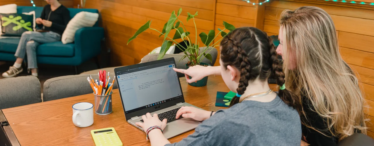 Two young women collaborate on a laptop at a wooden table with a calculator and pencils.
