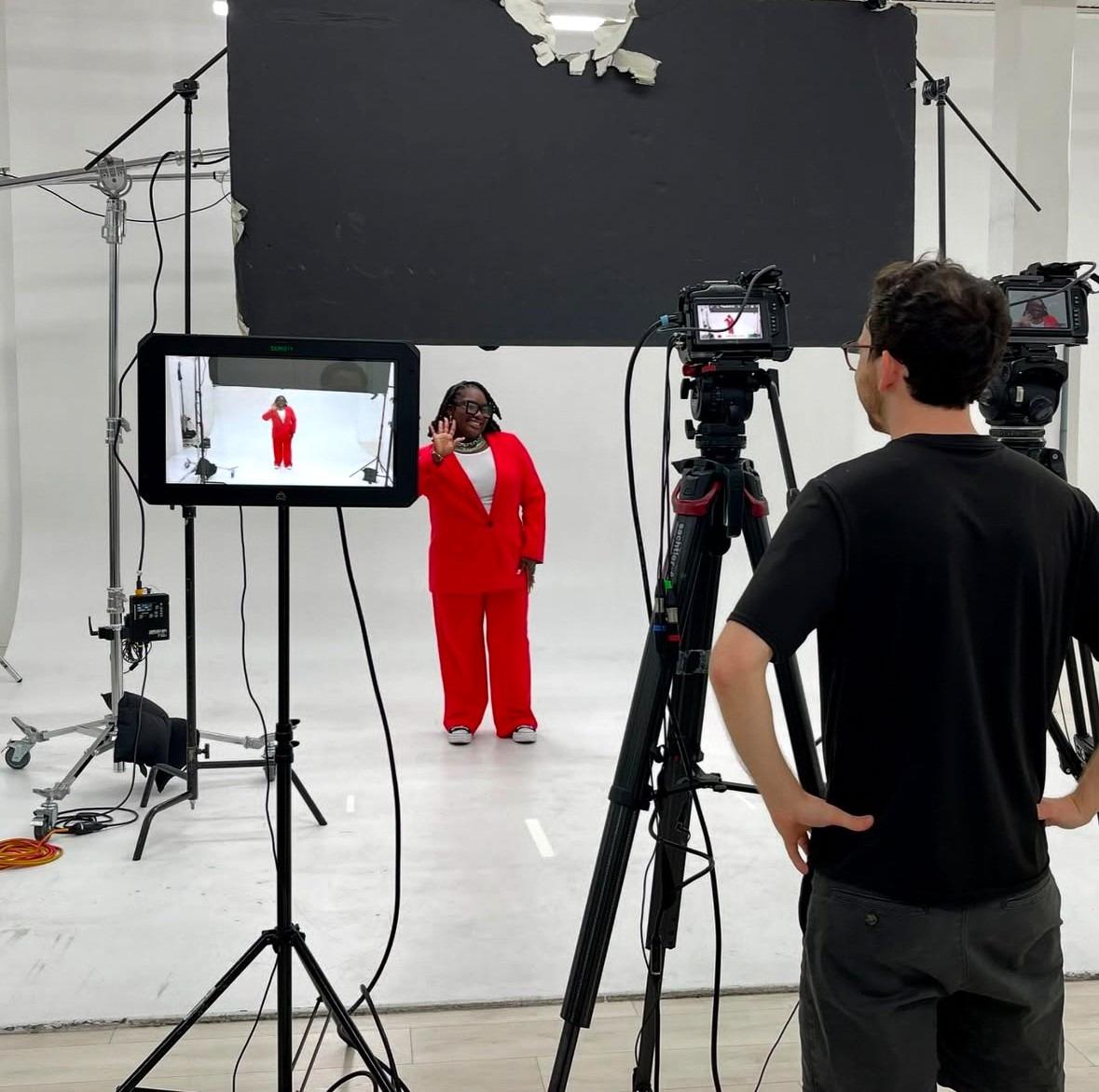 Une femme vêtue d’un costume rouge vif salue pendant qu’elle est filmée par une équipe de tournage dans un studio professionnel aux murs blancs.